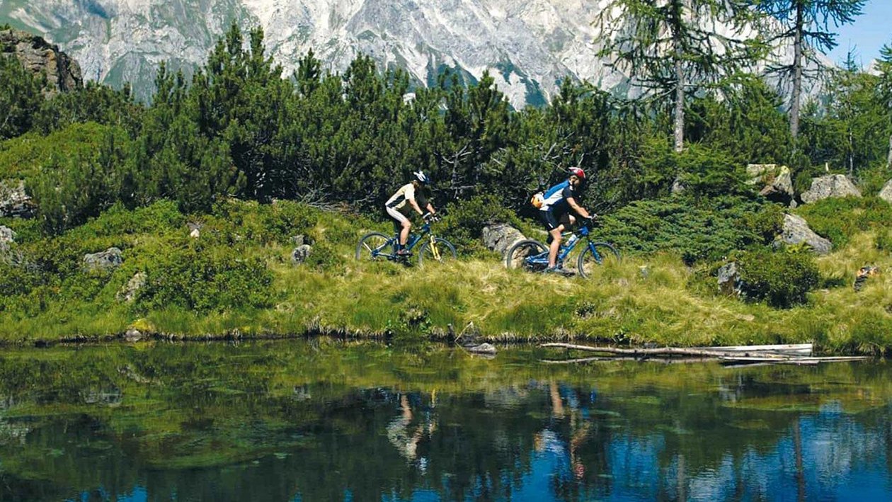 Abenteuer beim Mountainbiken Zwei Personen fahren mit Mountainbikes auf einem Weg entlang eines Teichs umgeben von Bergen.
