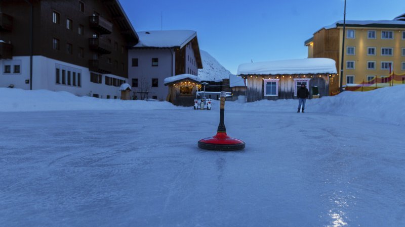Curling auf dem Eis im Winter Ein Curlingstein liegt auf einem Eislaufplatz umgeben von schneebedeckten Gebäuden bei Dämmerung.