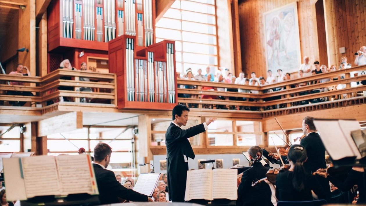 Orchesterperformance im Konzertsaal Dirigent leitet ein Orchester in einem voll besetzten Konzertsaal mit Publikum im Hintergrund.