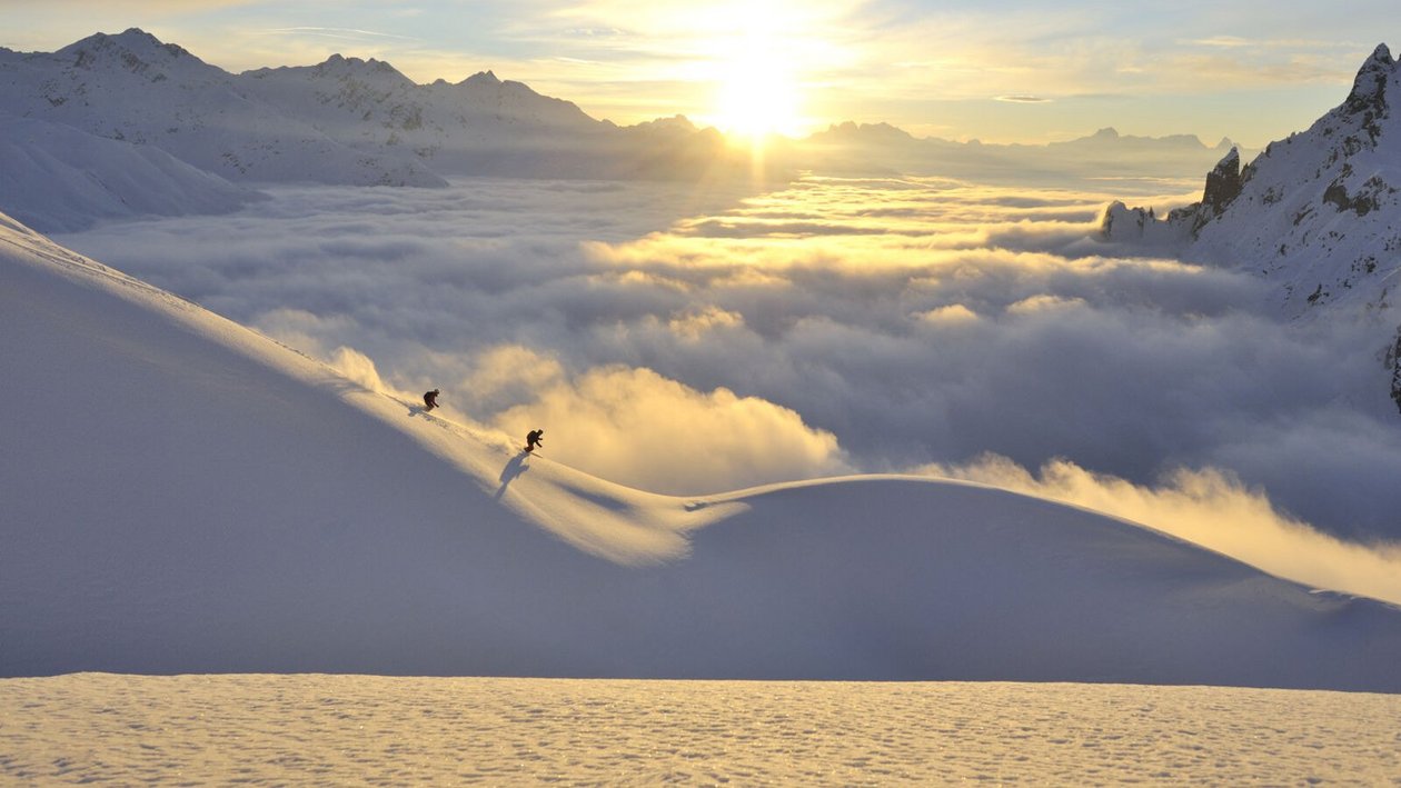 Schneelandschaft im Arlberg Zwei Skifahrer fahren über eine schneebedeckte Piste bei Sonnenaufgang über Wolken