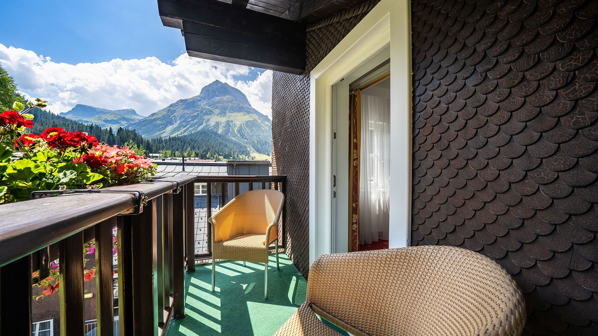 Balkon mit Bergblick im Tannbergerhof Zwei Rattanstühle auf einem Balkon mit Blumen und Blick auf die Berge in Lech, Österreich