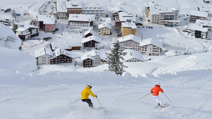 Skifahren im Herzen der Alpen Zwei Skifahrer fahren eine verschneite Piste in einem malerischen Bergdorf hinunter.