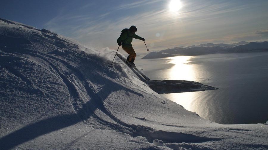 Freeride Abenteuer auf der Piste Skifahrer springt von schneebedecktem Hang mit Bergen im Hintergrund und Wasser.