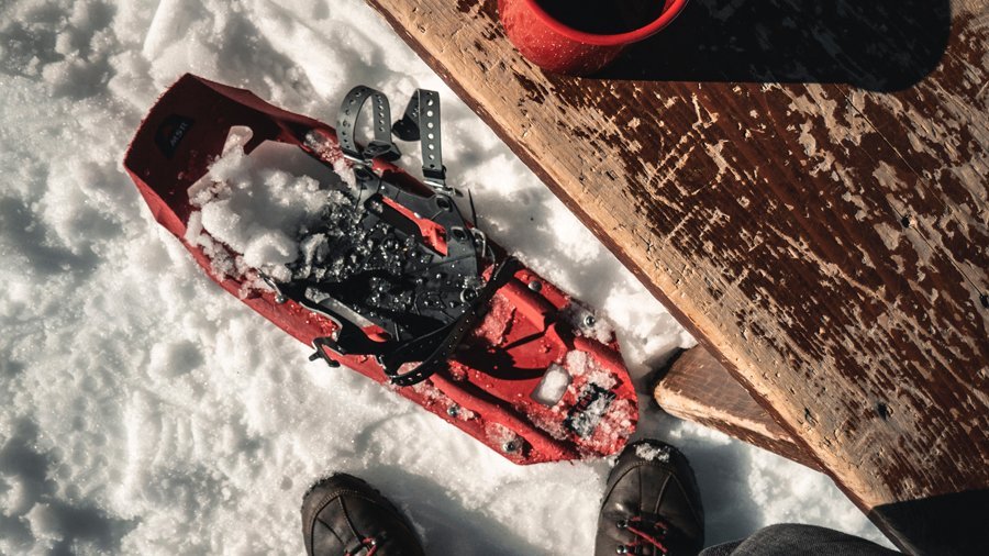 Schneeschuh Abenteuer in Arlberg Rote Schneeschuhe liegen im Schnee neben einer Holzbank und einer Tasse, Füße sind im Vordergrund sichtbar.