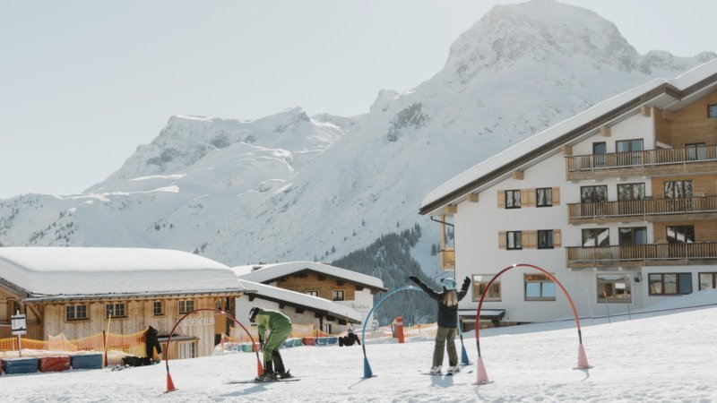 Skifahren im Winterparadies Zwei Skifahrer auf einer verschneiten Piste mit Bergen im Hintergrund und einem nahegelegenen Skigebäude.