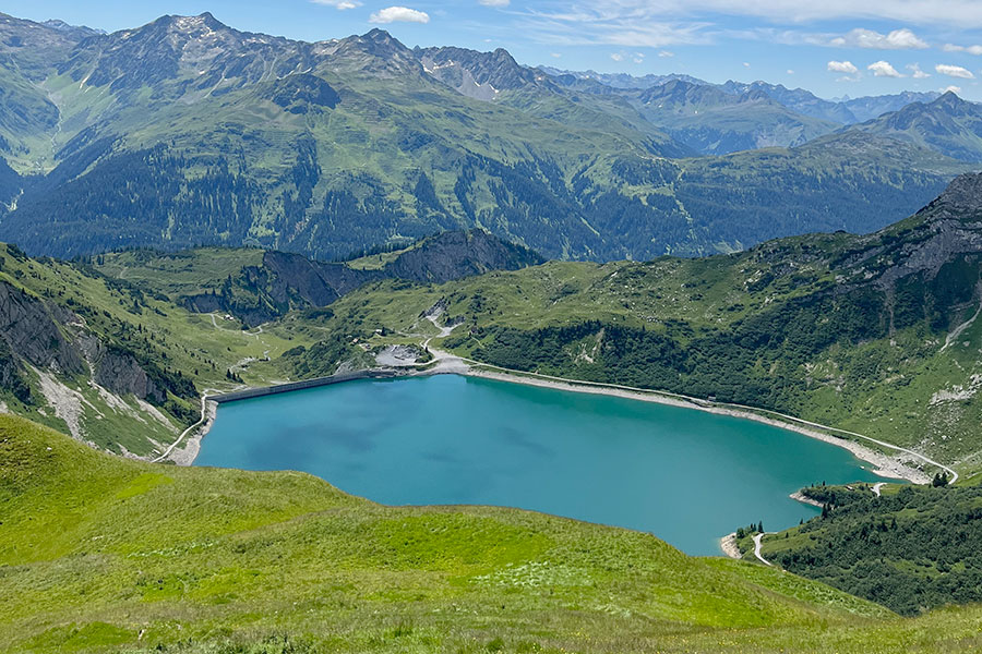 Panorama des Spullersee Bergsee mit türkisblauem Wasser am Arlberg, umgeben von grünen Wiesen und der Vorarlberger Gebirgskulisse