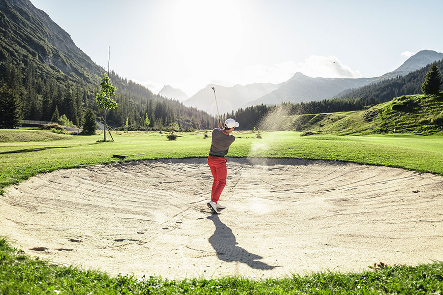 Perfekter Golfschwung in den Bergen Golfer spielt einen Schlag aus dem Bunker auf einem Golfplatz am Arlberg mit Bergkulisse im Hintergrund