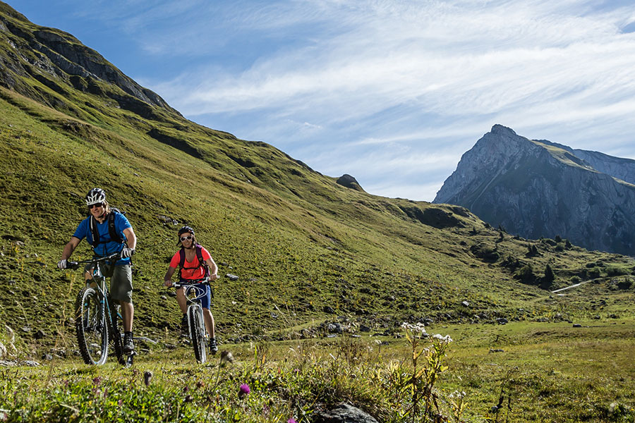 Abenteuer beim Mountainbiken in den Alpen Zwei Mountainbiker auf einem alpinen Trail am Arlberg mit grünen Wiesen und Bergkulisse im Hintergrund © Hanno Mackowitz