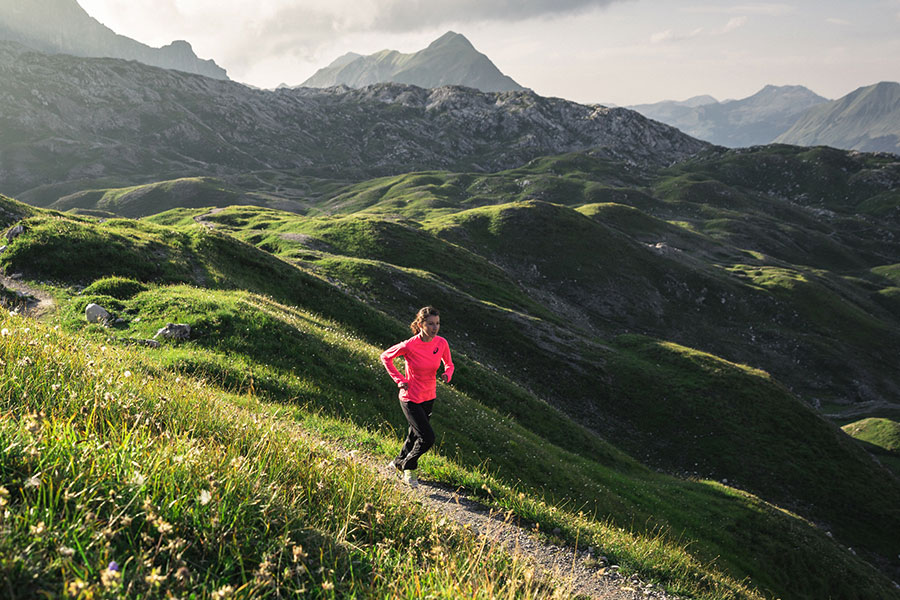 Aktiv im Gebirge Trailrunnerin in pinkem Shirt läuft auf einem schmalen Pfad durch die hügelige Berglandschaft am Arlberg © Hanno Mackowitz