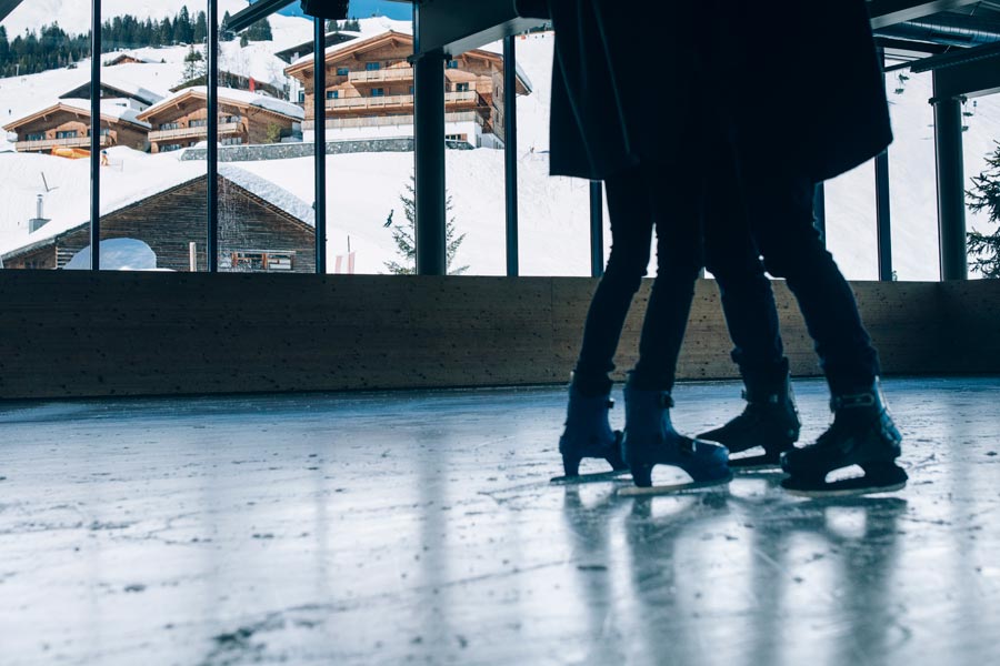 Romantisches Eislaufen im Winterzauber Eislaufbahn mit Blick auf verschneite Chalets in Lech, zwei Personen in Schlittschuhen stehen einander gegenüber © Daniel Zangerl