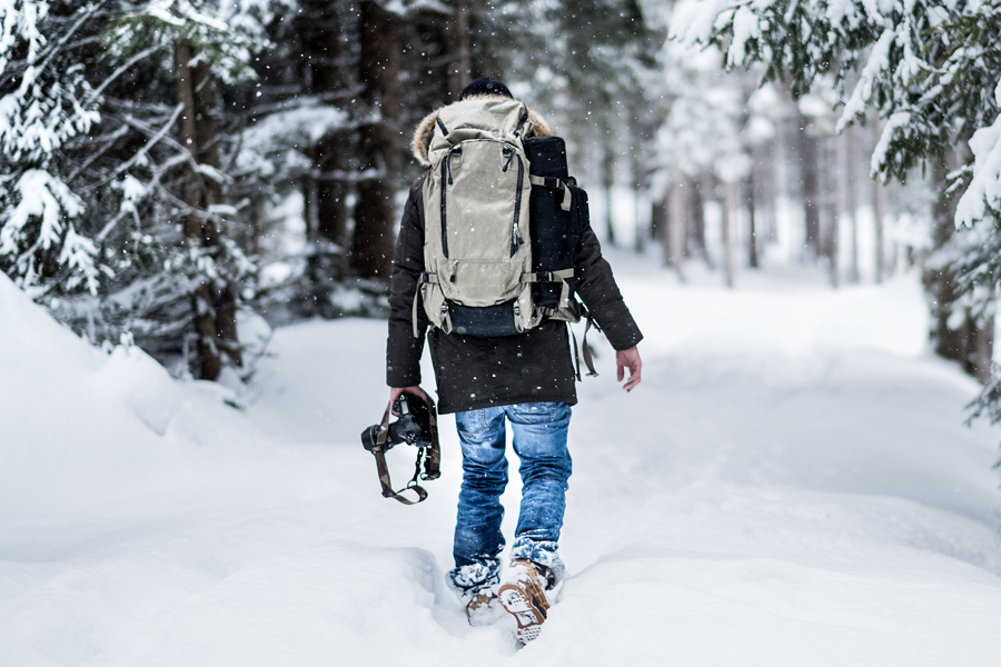 Wunderschöne Fotomotive in der Winterlandschaft in Lech am Arlberg Mann mit Rucksack geht auf einem verschneiten Pfad Im wald spazieren, in der linken Hand befindet sich eine Kamera.