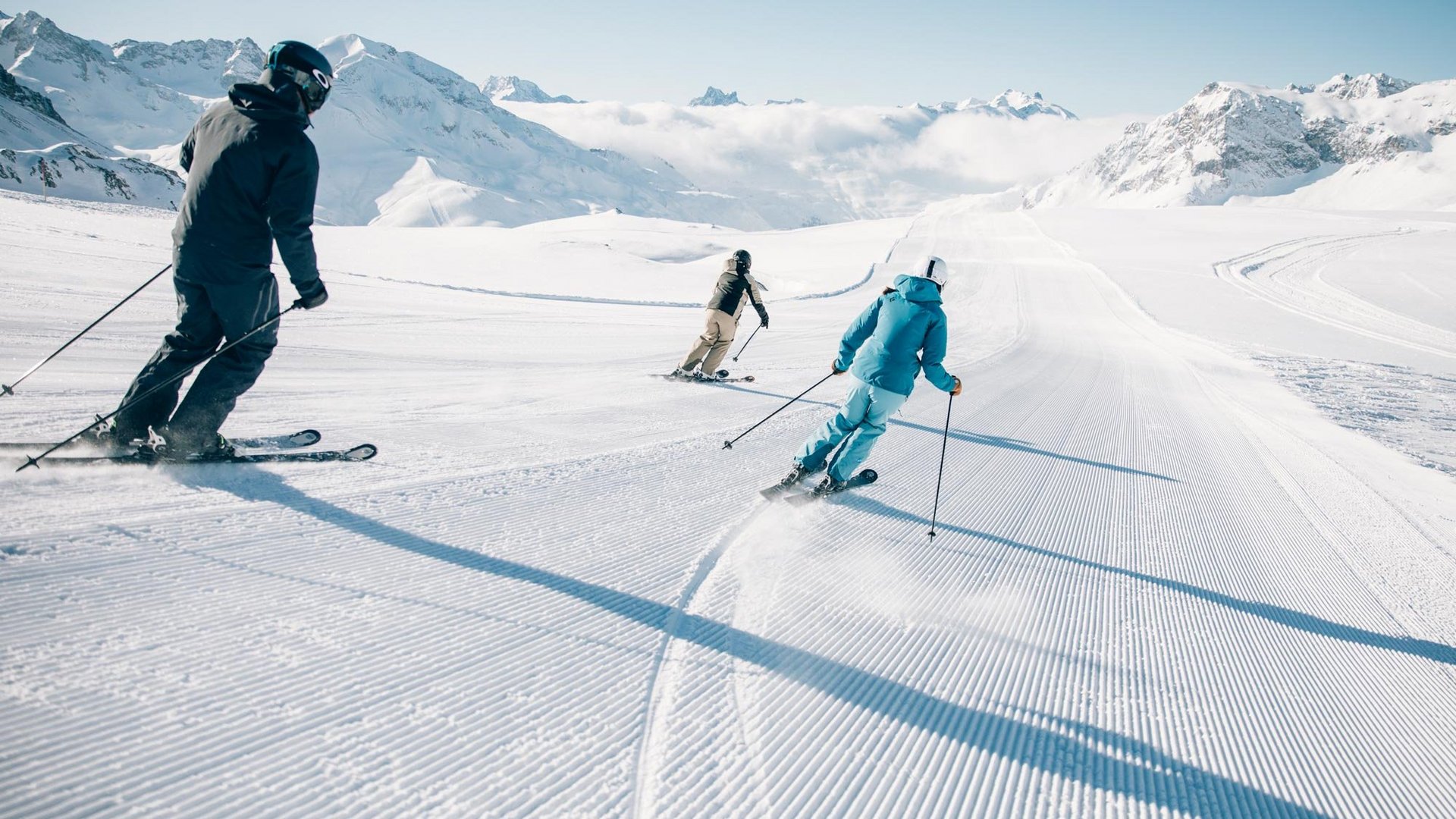 Schneebedeckte Pisten erobern Drei Skifahrer fahren eine schneebedeckte Abfahrt in den Bergen unter blauem Himmel.