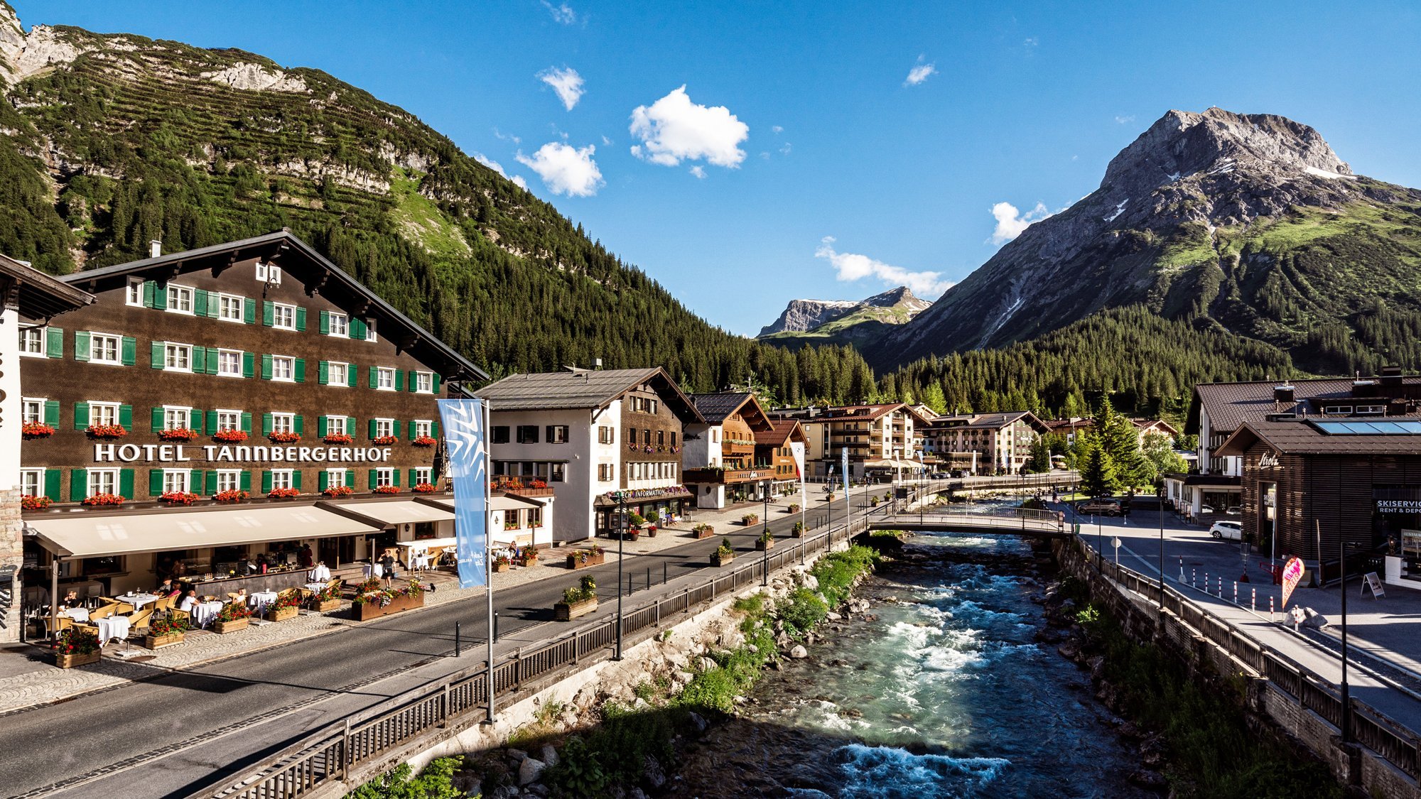 Das Hotel Tannbergerhof in der Außenansicht in Lech am Arlberg mit Gebirgsbach und Bergen im Hintergrund Malersches Alpendorf mit Hotel und Blick auf Berge und Flusslauf in der Naturkullisse.